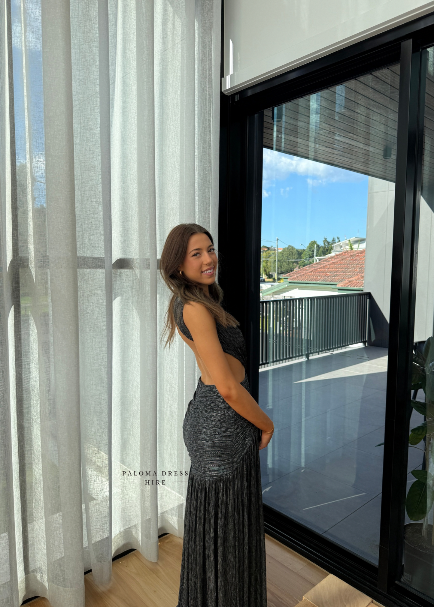 Woman in a gray dress standing by a large window with a view of a rooftop garden.