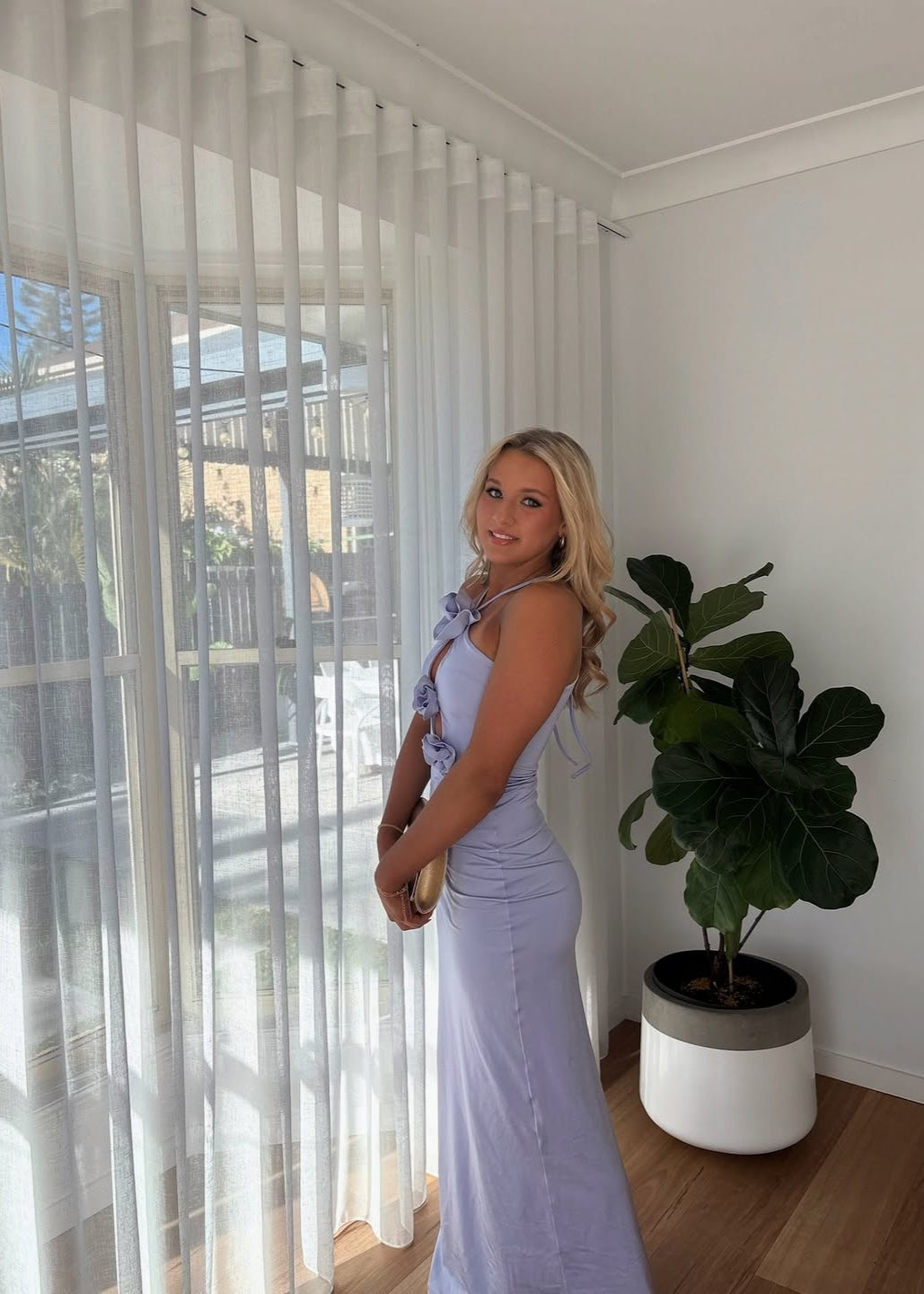 Woman in a light purple dress standing by a large window with a plant nearby.