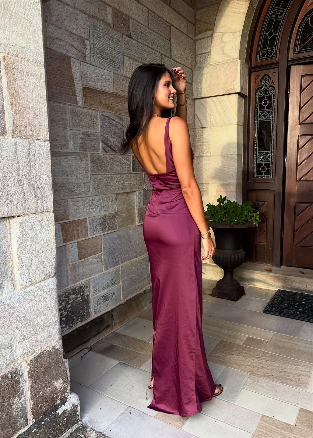 Woman in a burgundy evening gown standing in front of a stone wall and wooden door.
