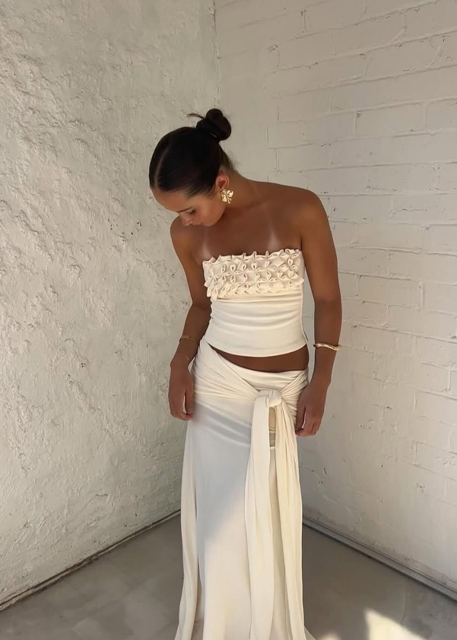 Woman wearing a white strapless top and flowing white skirt against a white brick wall.