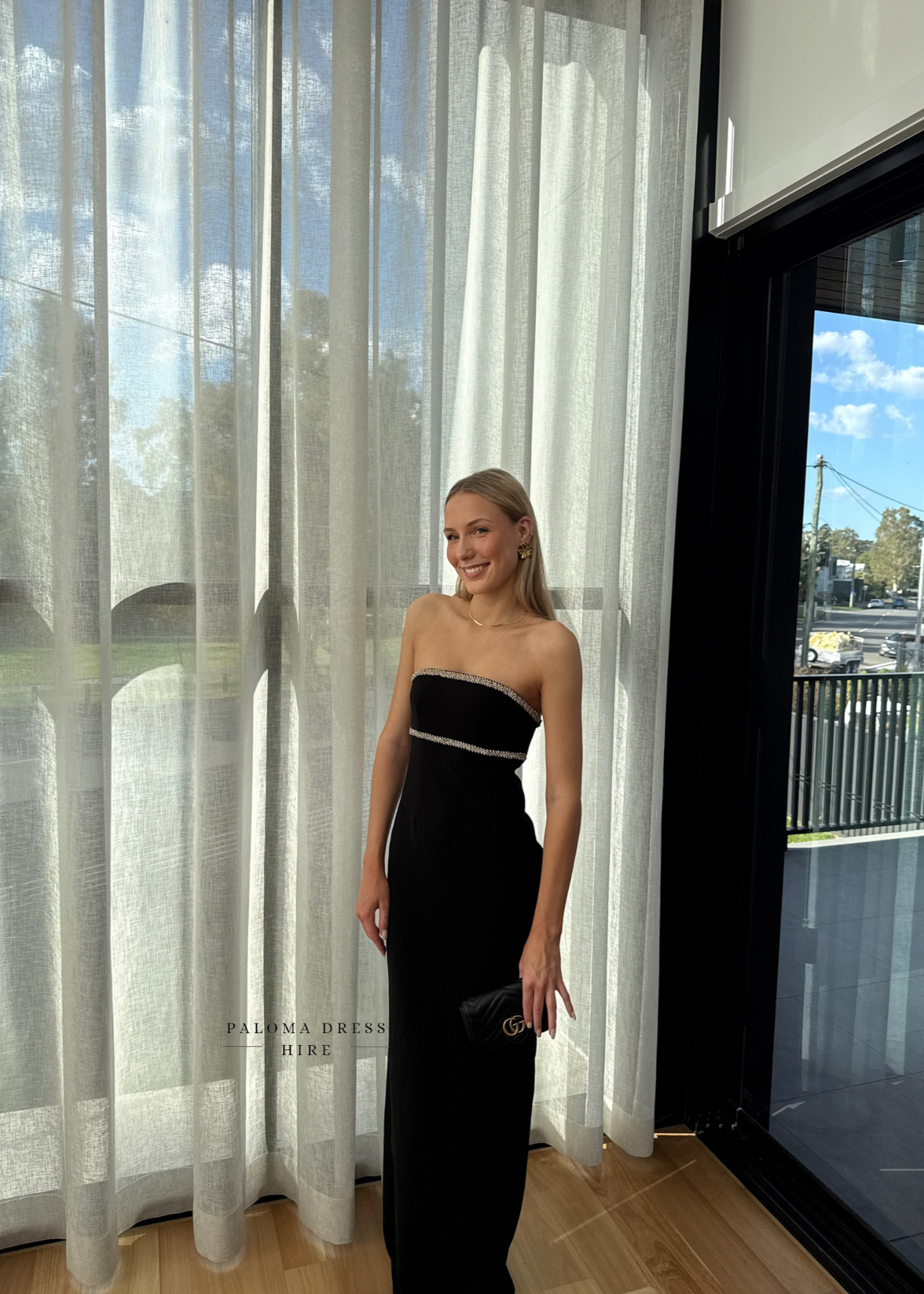 Woman in a black strapless dress standing in front of sheer curtains with 'Palmia Dress Hire' branding.