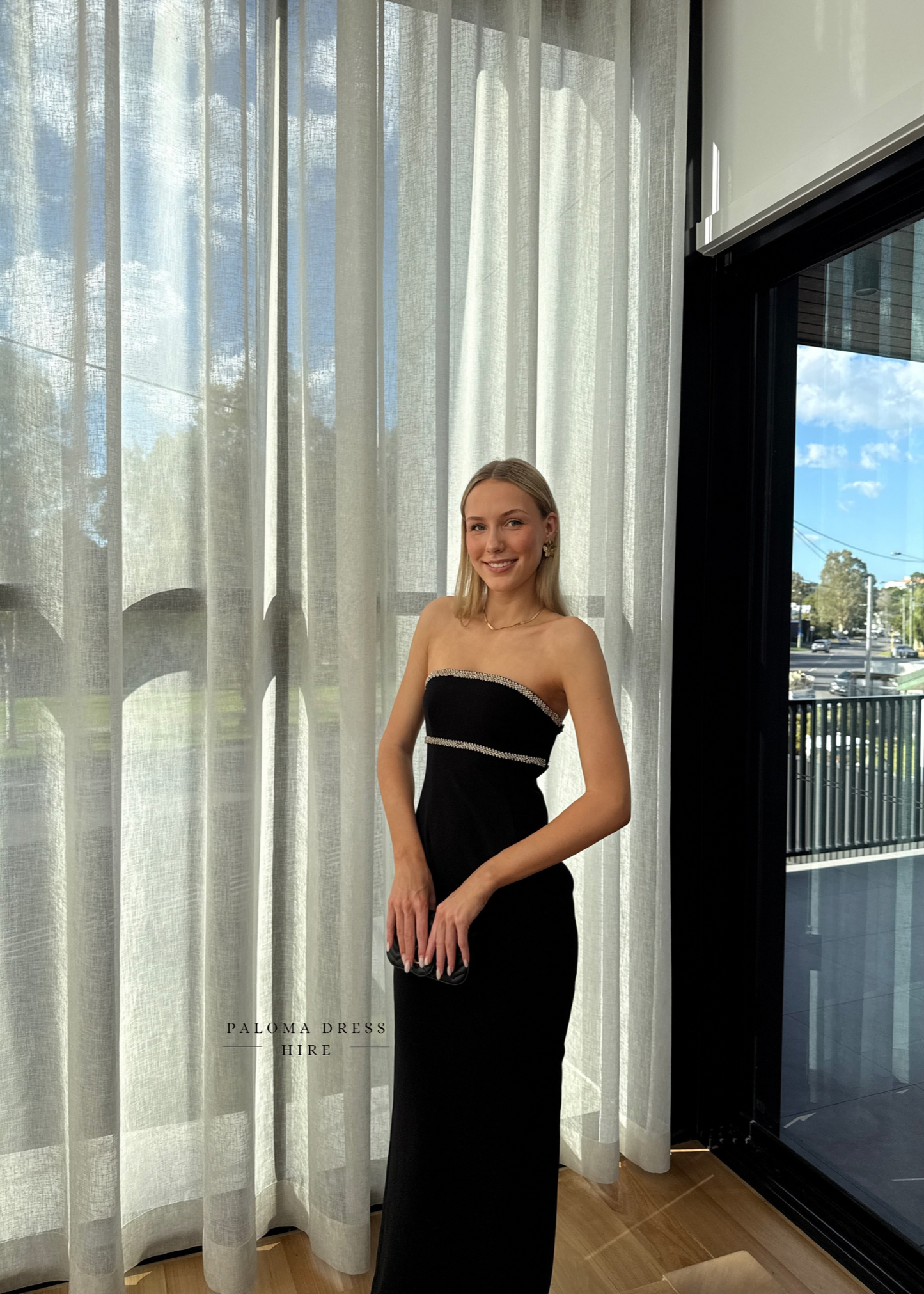 Woman in a black strapless dress standing in front of sheer curtains with a cityscape view.
