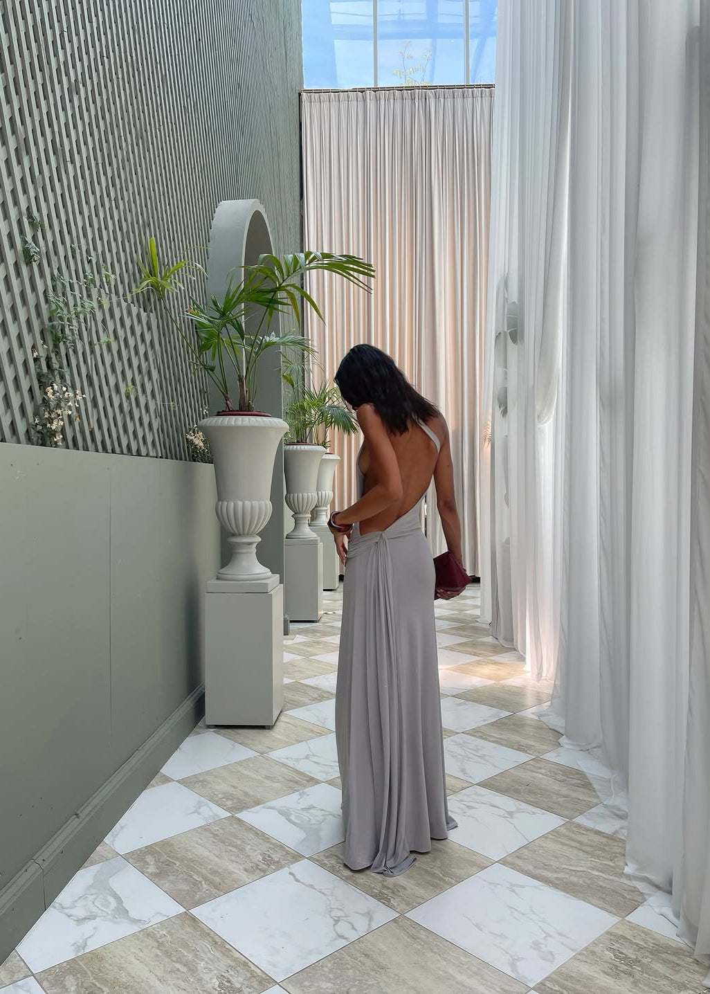 Woman in a long, light-colored dress standing in a modern indoor setting with plants and large windows.