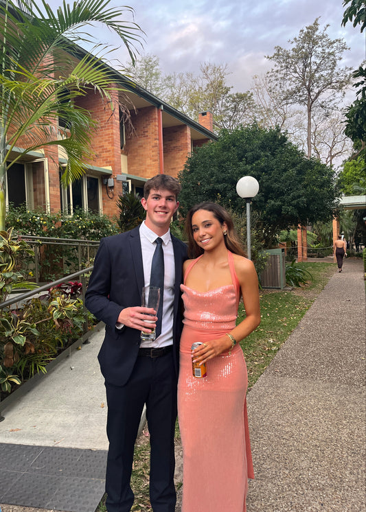 Two people in formal attire standing outdoors with a house and garden in the background.