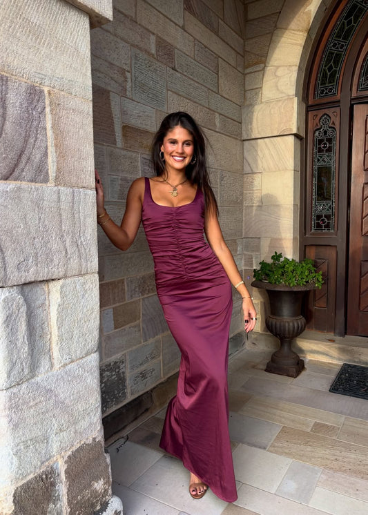 Woman in a purple dress standing against a stone wall with a wooden door in the background.