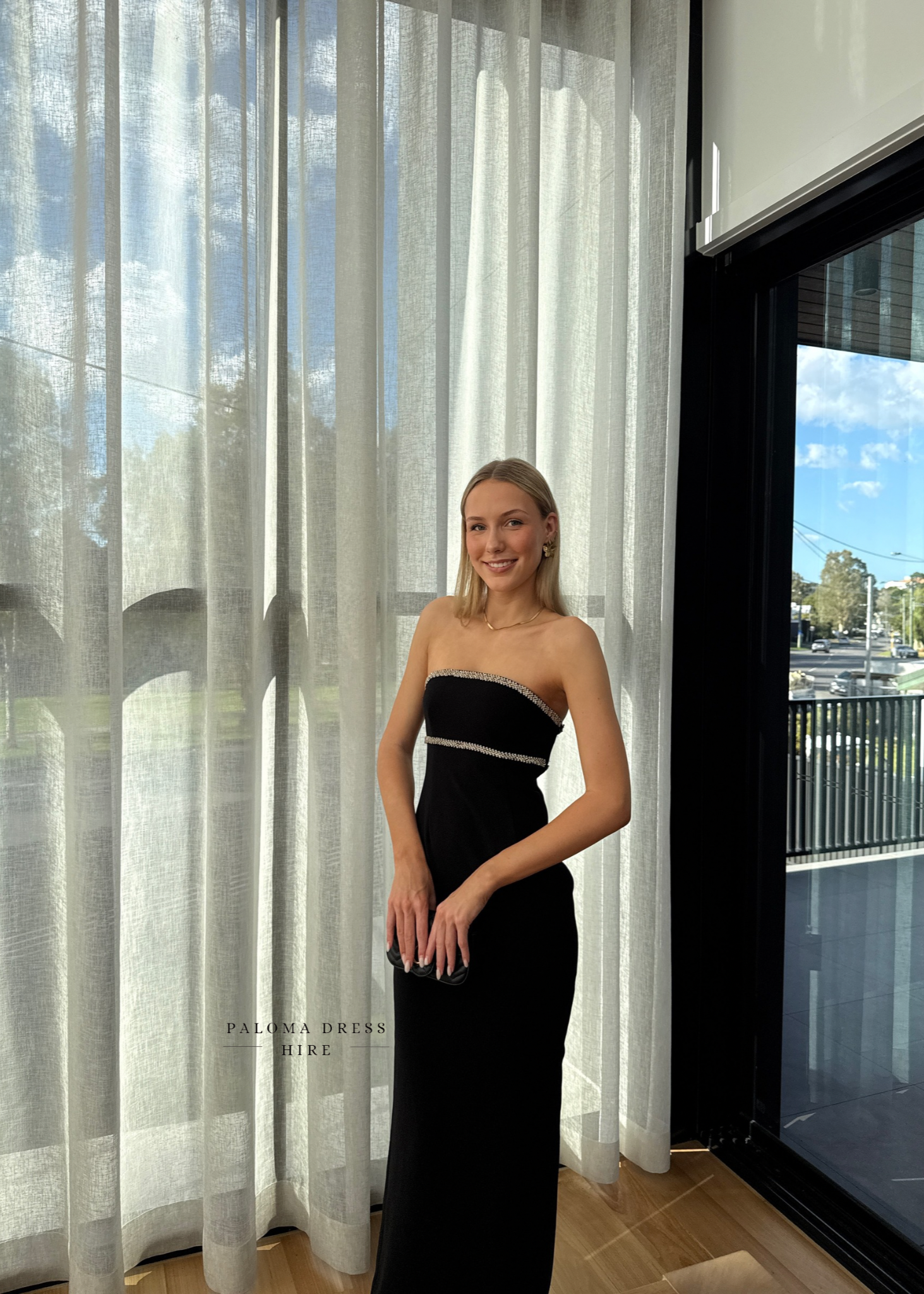Woman in a black strapless dress standing in front of sheer curtains with a cityscape view.