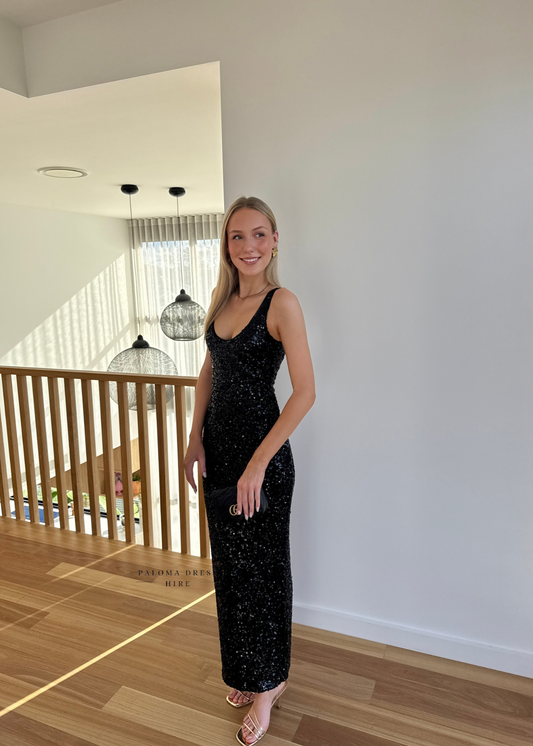Woman in a black sequin dress standing in a room with wooden flooring and a white wall.