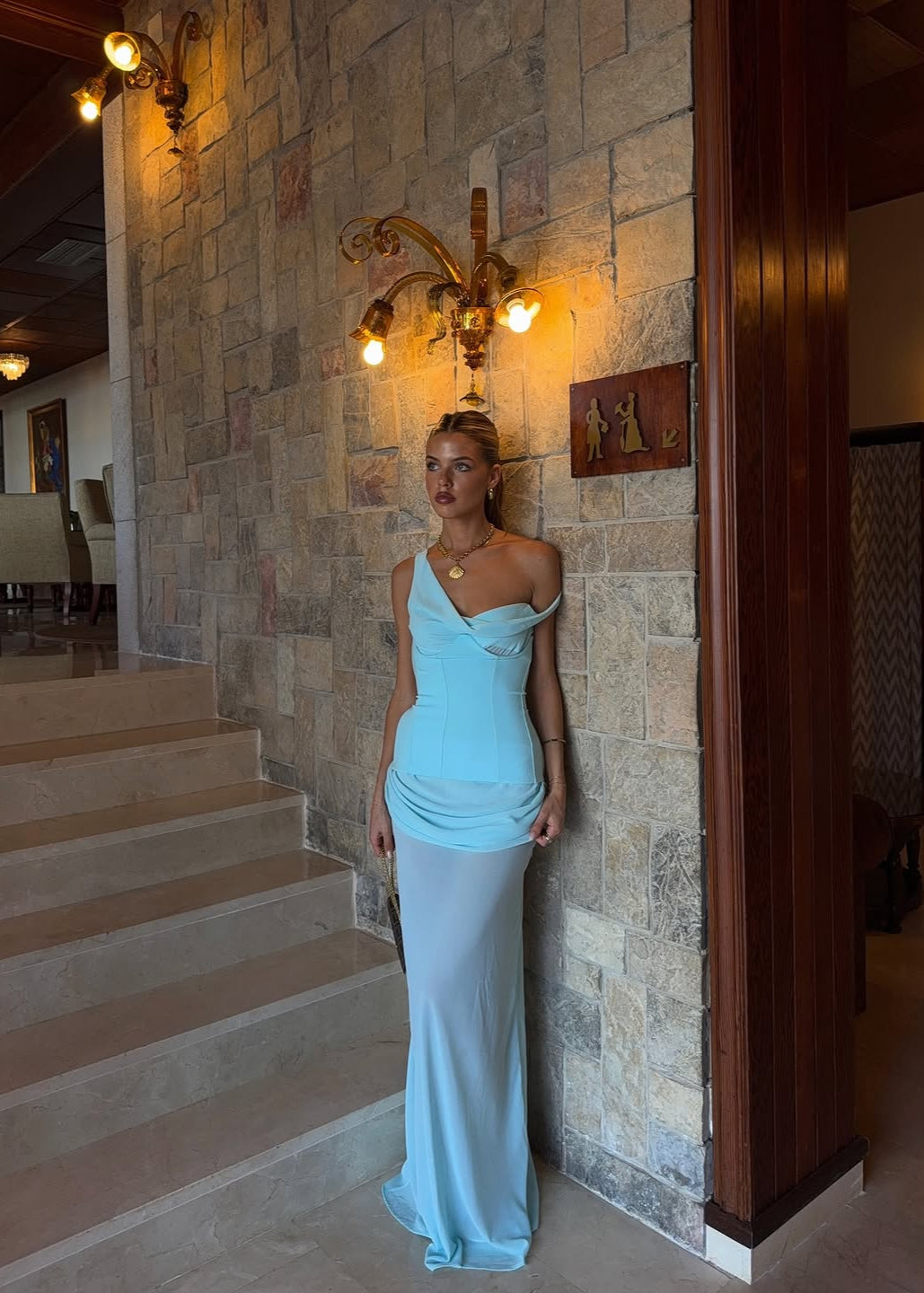 Woman in a light blue dress standing in a room with stone walls and wooden beams.